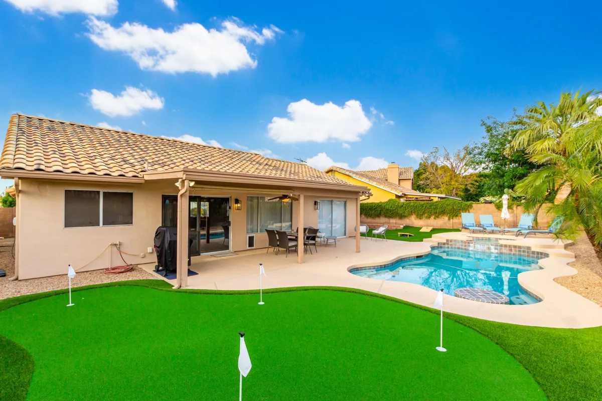 A sunlit scene of a backyard with a sparkling swimming pool adjacent to a lush putting green and a comfortable patio seating area. The image also captures a well-manicured lawn, offering a sense of openness and inviting outdoor living.