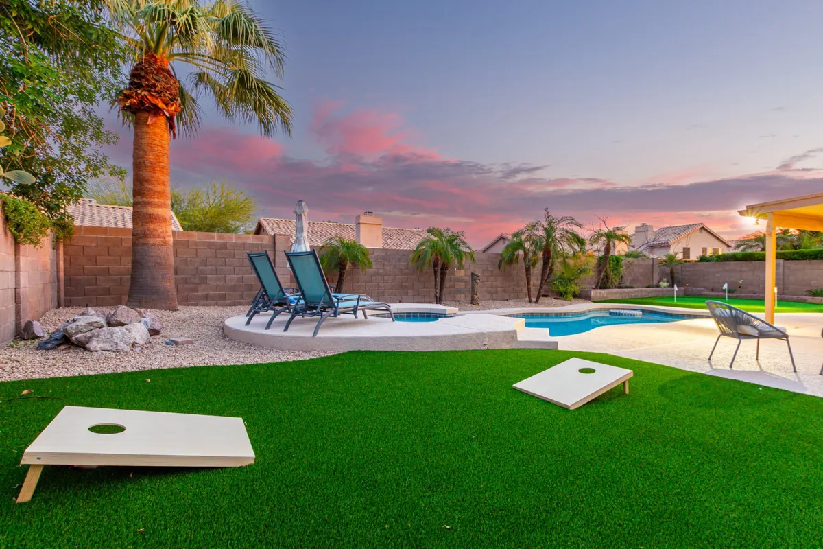 A lively backyard setting featuring a swimming pool with lounge chairs and a cornhole game set up on the grass. The space is designed for casual enjoyment, with activities for all ages and plenty of room for sunbathing and outdoor games.