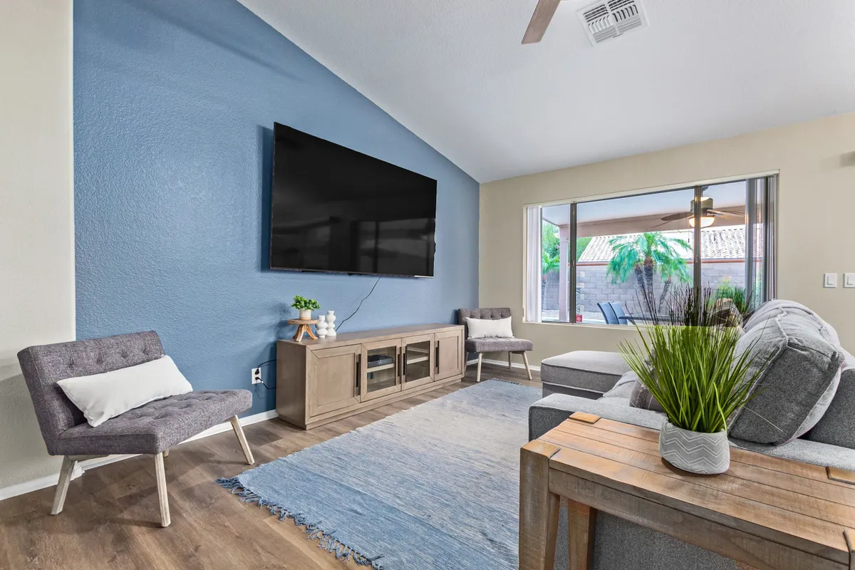 A contemporary living room with a large flat-screen TV as the focal point, surrounded by a plush sofa seating area, a stylish armchair, a wooden coffee table, and a decorative plant adding a touch of greenery. Bright natural light streams through the windows, and the room feels both comfortable and modern—ideal for family gatherings or casual movie nights.