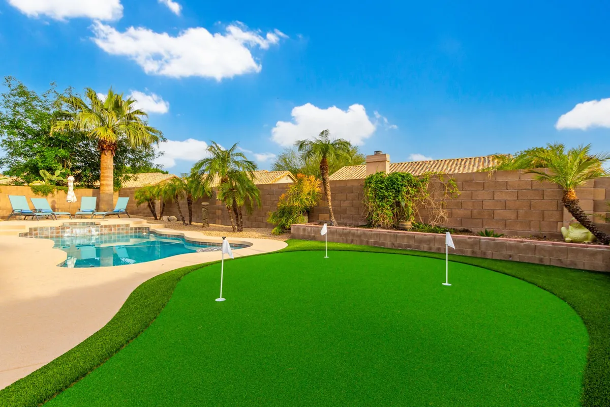 This image shows a vibrant backyard with a sparkling swimming pool, lush green putting green, palm trees, and several lounge chairs. The scene is inviting and playful, with ample space for sunbathing, swimming, and practicing golf, all set against the backdrop of a sunny Mesa afternoon.