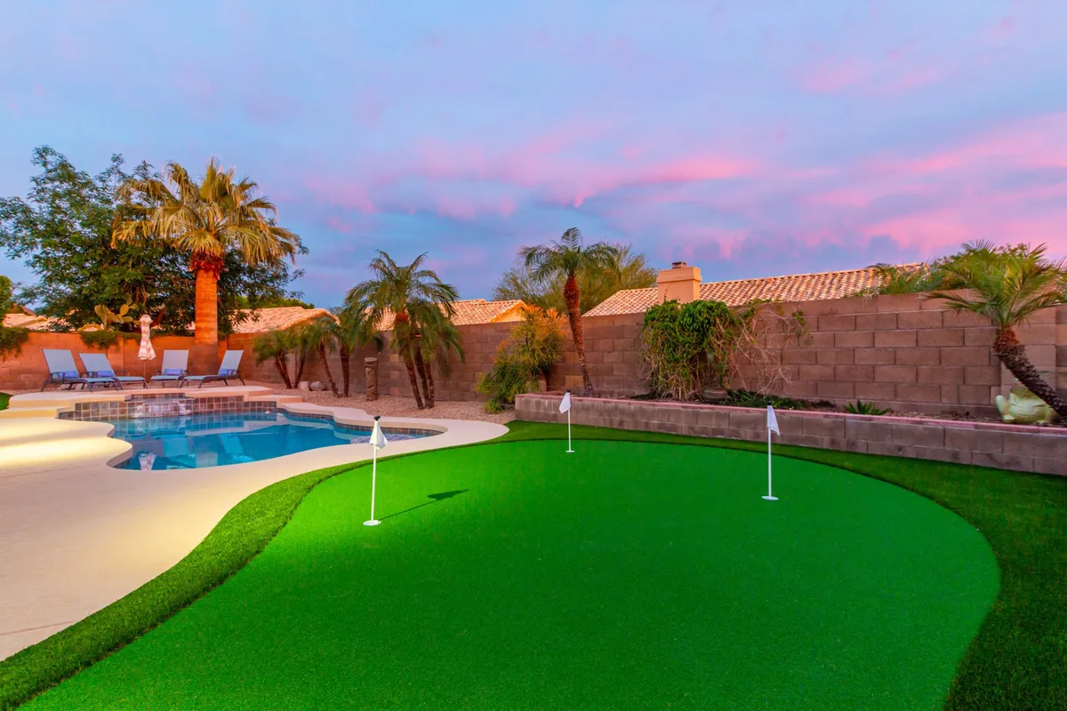 This image showcases a tranquil backyard setting featuring a sparkling swimming pool, lush putting green, swaying palm trees, and a colorful sky at sunset. The pool and putting green are the stars, with ample space for relaxation and play, embodying a true resort-style escape.
