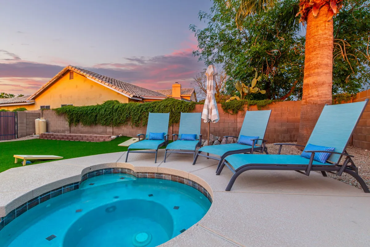 This image centers on a backyard hot tub surrounded by plush lounge chairs and a sun umbrella, all set on green grass amidst thoughtful landscaping. The scene is peaceful and rejuvenating, ideal for relaxation and socializing, with the setting sun casting a warm glow over the space.