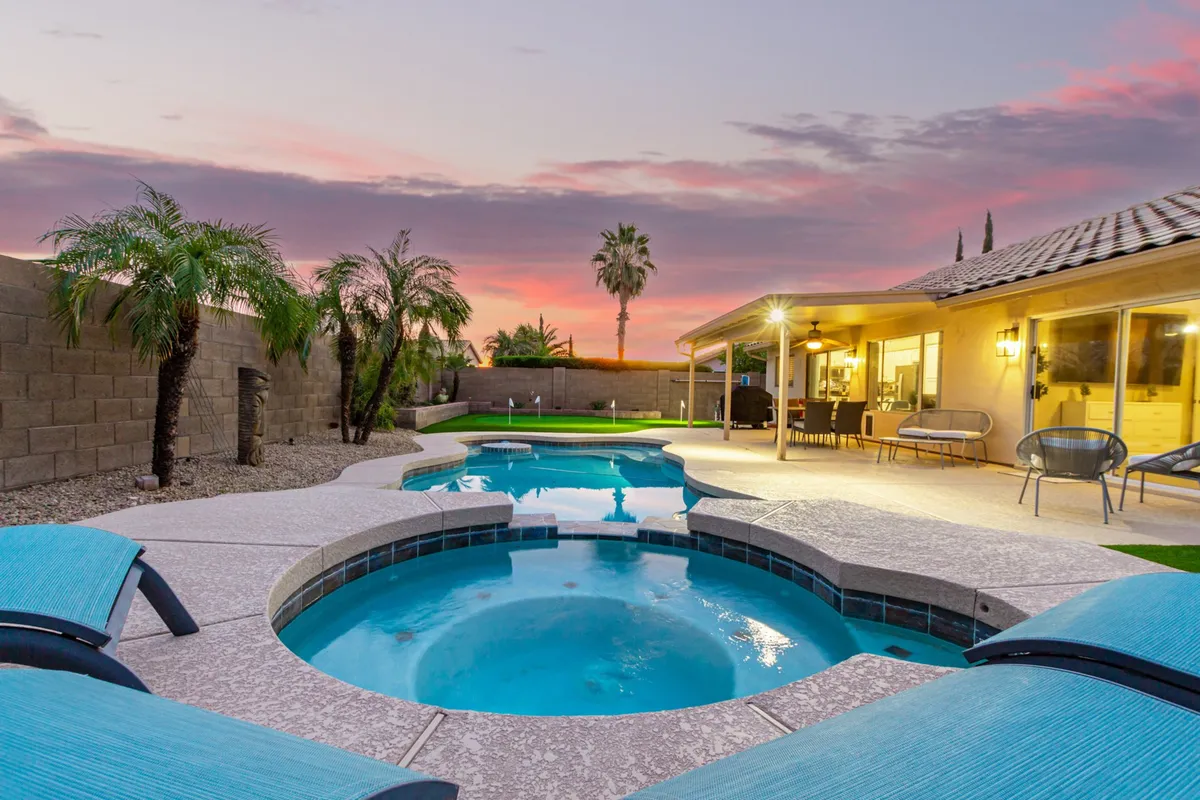 The image showcases a serene backyard with a sparkling swimming pool, an inviting jacuzzi, and a comfortable outdoor seating area. The landscaping is lush and well-maintained, with palm trees and decorative lighting enhancing the tranquil atmosphere. A dramatic sunset sky completes the luxurious, resort-style setting.
