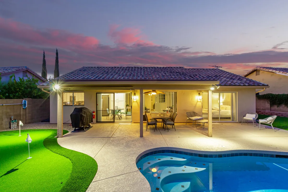 This image features a vibrant backyard with a swimming pool, a spacious outdoor seating area, a green lawn, and a dining table set for outdoor meals. The scene captures a sunny, cheerful energy and emphasizes space for gatherings and relaxation.