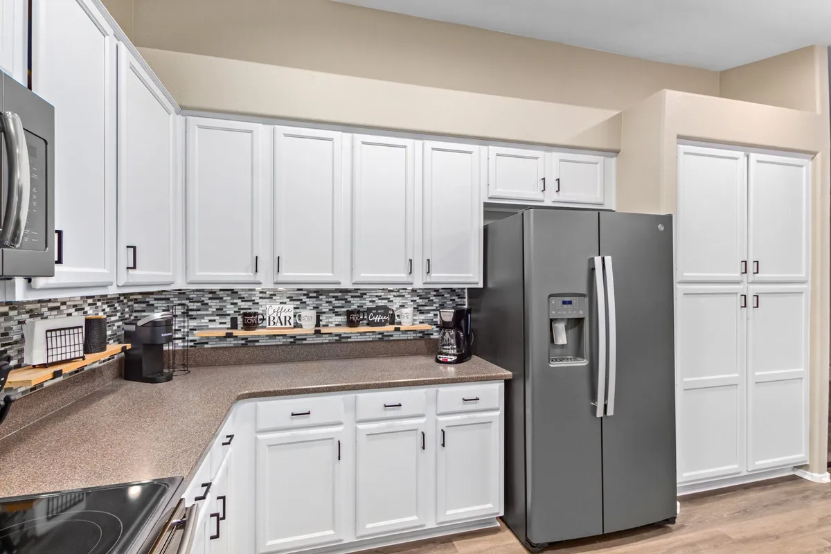 A close-up view of the kitchen area with white cabinets, a decorative mosaic tile backsplash, stainless steel refrigerator, and a coffee maker on the counter. The inviting setup emphasizes morning rituals and casual meal prep.