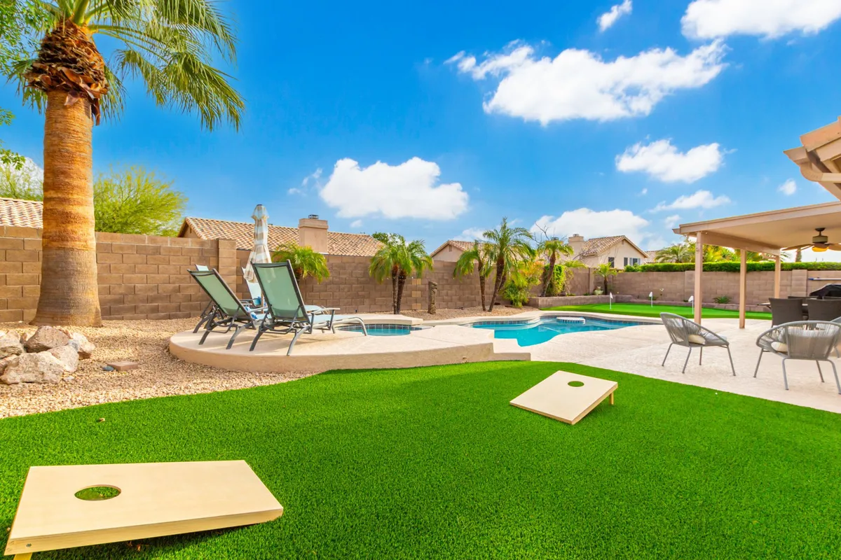 This image captures a vibrant backyard setup with a sparkling swimming pool, lawn area featuring cornhole boards, patio seating, and elegant palm trees. The scene is bright and lively, reflecting both recreational and social activities.