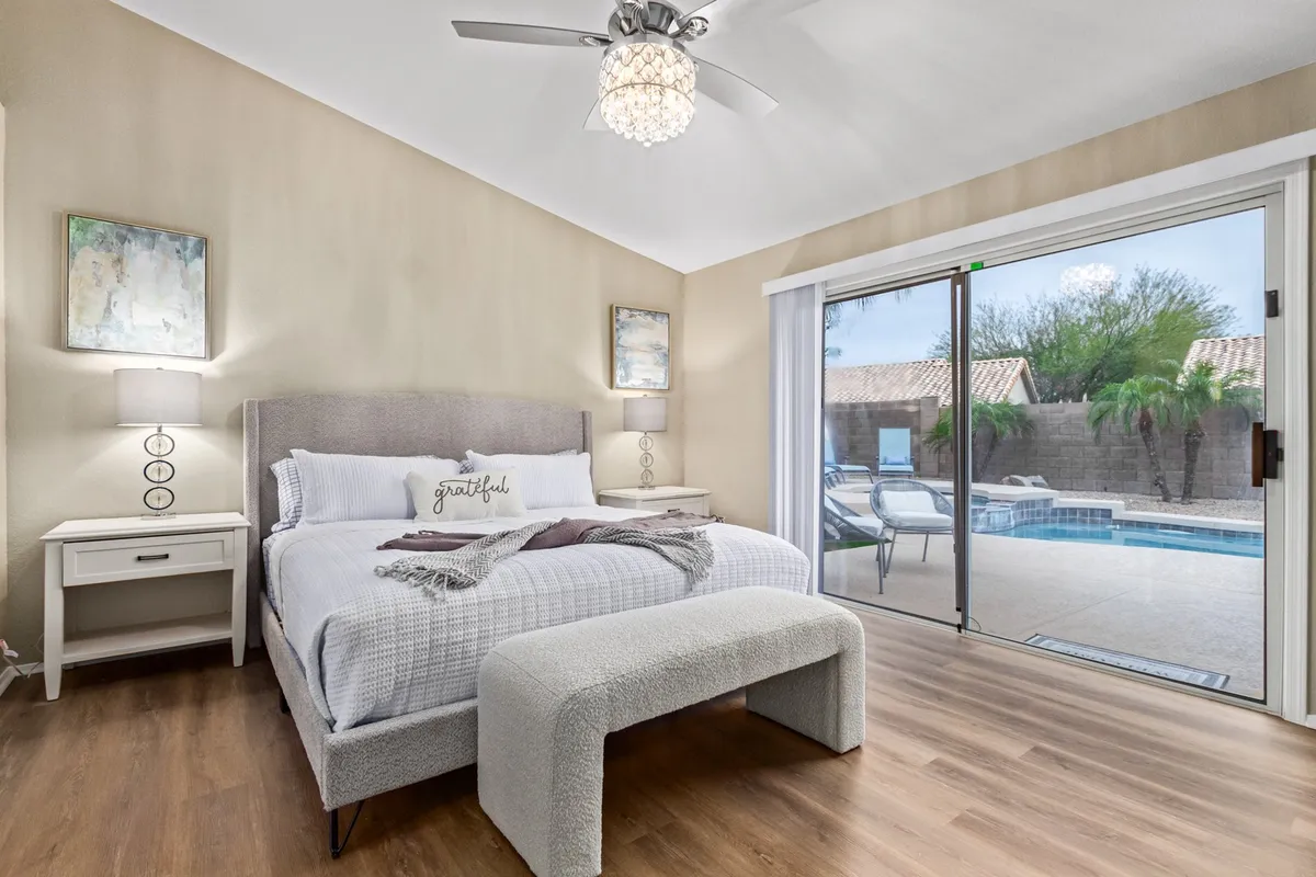 A serene main suite featuring a neatly made bed with decorative pillows, elegant artwork on the walls, and a sliding glass door that opens directly to the pool area. Soft natural light enhances the atmosphere, and the pool is visible just outside, hinting at easy outdoor access.
