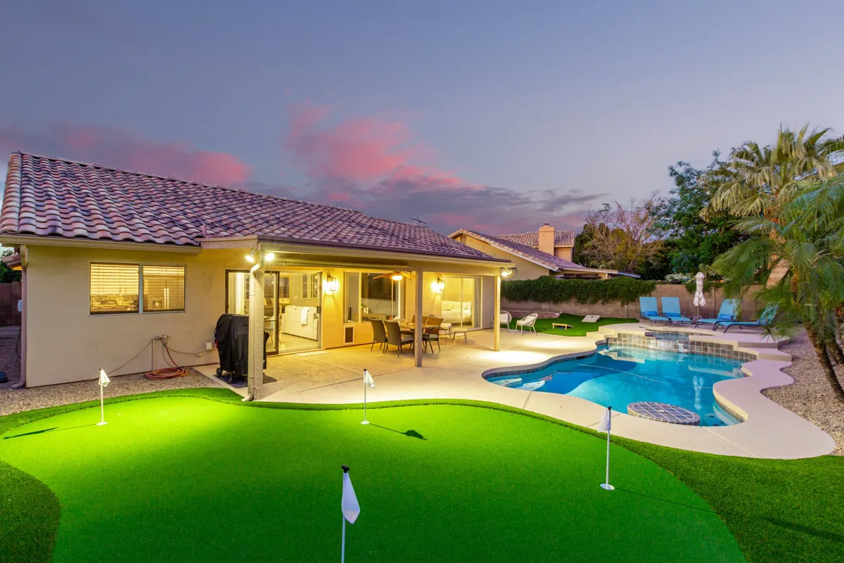 The image showcases the home's stunning backyard oasis in Mesa, Arizona, featuring a sparkling swimming pool surrounded by comfortable outdoor seating, a manicured putting green for golf practice, and lush landscaping perfect for sunset gatherings.